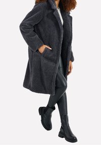 Donna che indossa un lungo cappotto grigio scuro a trama, pantaloni in pelle neri, stivali chunky neri e una maglietta bianca, in piedi con una mano nella tasca del cappotto.