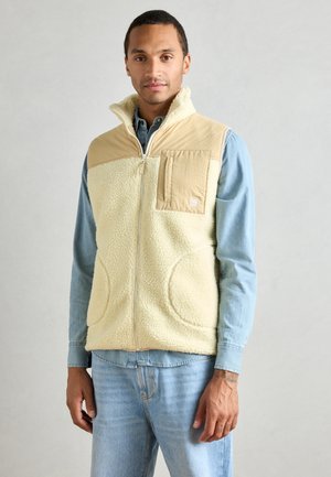Fleece vest in cream with tan top panel and pocket. Features zip closure, side pockets, and a high collar. Worn over a denim shirt.
