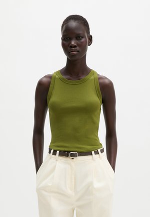 Femme portant un haut sans manches vert olive rentré dans un pantalon blanc taille haute avec une ceinture noire, debout les mains dans les poches.