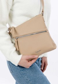 Krémová kožená crossbody taška s texturovaným povrchem, která má stříbrný zipový uzávěr a uzlový detail na popruhu.