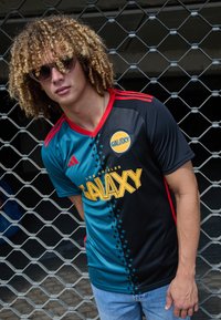 Camiseta de fútbol negra y verde azulado con acentos rojos y un gran logo amarillo "Galaxy". Presenta un patrón de estrellas a lo largo del costado. Se lleva con unos jeans azul claro.