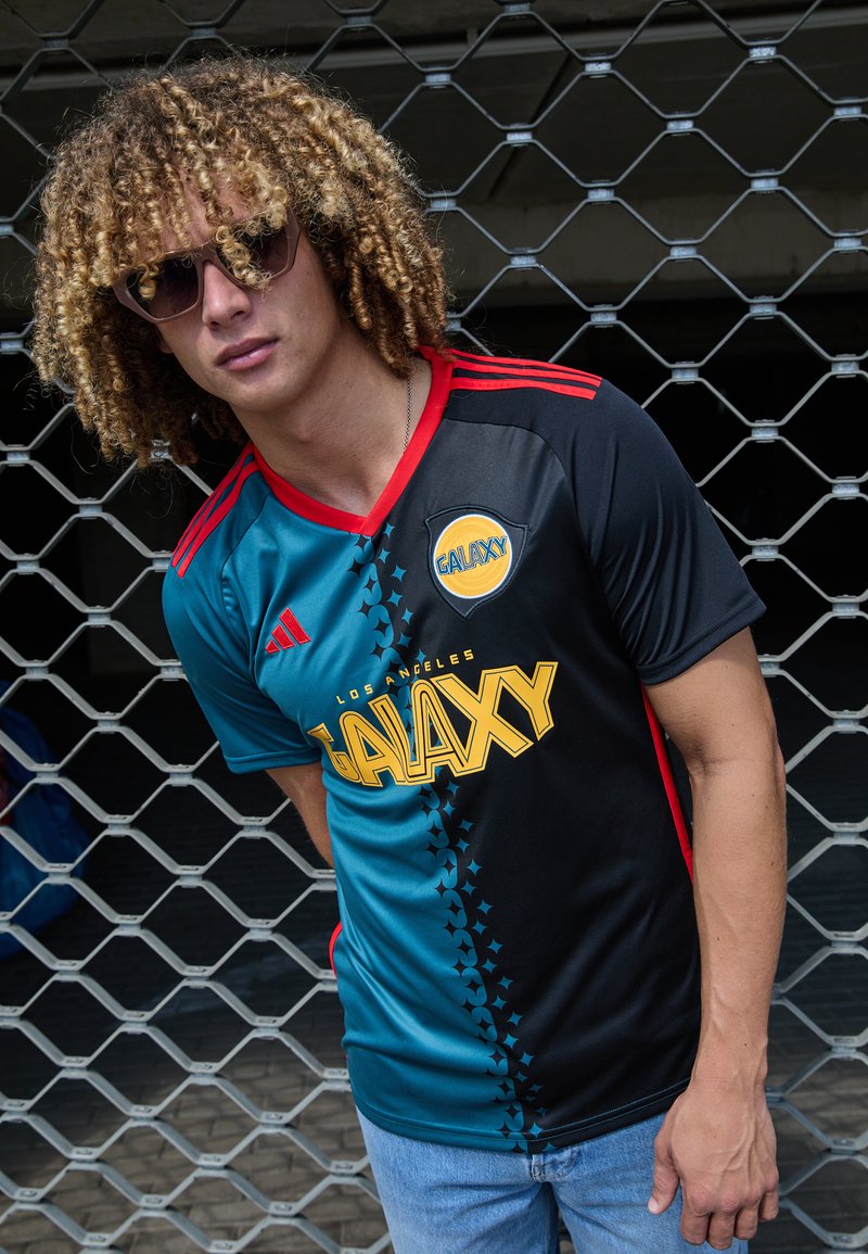 Camiseta de fútbol negra y verde azulado con acentos rojos y un gran logo amarillo "Galaxy". Presenta un patrón de estrellas a lo largo del costado. Se lleva con unos jeans azul claro.