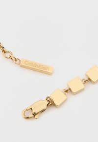 Χρυσόχρωμο βραχιόλι αλυσίδας με τετράγωνα κλειδιά, κούμπωμα και εγχαραγμένη ορθογώνια ετικέτα που αναγράφει "Calvin Klein". Λείος υφασμάτινος.