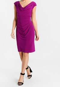 Abito fucsia con maniche a cappuccio, scollatura a foulard e texture arricciata. Abbinato a sandali neri con cinturino alla caviglia. Tessuto liscio, design fino al ginocchio.