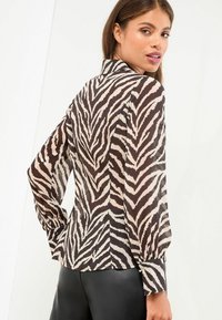 Doorzichtig bloesje met een zebrapatroon in tinten bruin en crème. Het heeft lange mouwen en een kraag met details aan de manchetten.