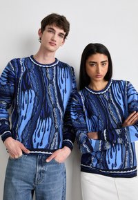 Pulls en tricot dans différentes teintes de bleu avec des motifs en vagues, dotés de poignets et d'un ourlet côtelés, associés à un jean clair et une jupe blanche.