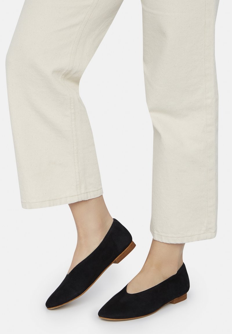 Pieds portant des ballerines pointues en daim noir avec des semelles camel, associées à un pantalon court écru sur fond blanc.