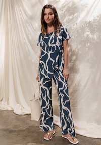 Haut bleu marine et pantalon ample avec motif abstrait blanc, en tissu lisse. Le mannequin tient un sac beige et porte des tongs blanches.