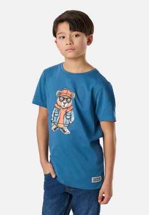 Dreng med kort mørkt hår iført blå grafisk t-shirt med bjørn i hovedtelefoner og jeans, stående med den ene hånd i lommen.