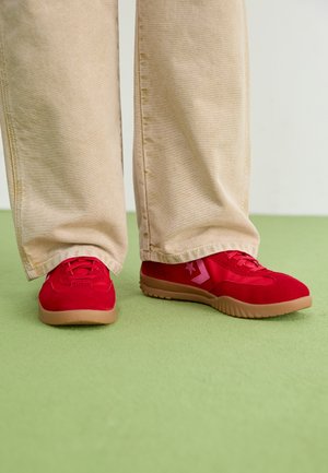 Rote Wildleder-Sneaker mit einer flachen Gummisohle und pinken Akzenten, kombiniert mit beigen, weit geschnittenen Hosen auf einem grünen Boden.