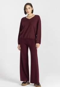 Kobieta stojąca w burgundowym swetrze z dekoltem w serek i dopasowanych szerokich spodniach, w brązowych butach na platformie, na białym tle.