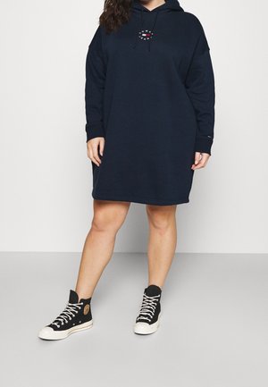 Kvinde iført en marineblå hættetrøjekjole med et lille rundt logo på brystet og sorte højtstående lærredssneakers med hvide snørebånd.