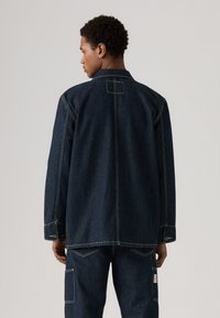 Mørk denimjakke med en avslappet passform, med spiss krage, knappedetaljer på ermene og kontrastsøm på ryggen og skuldrene.