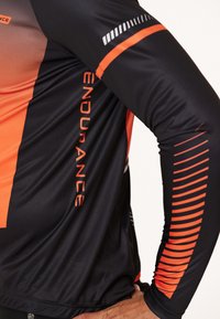 Svart och orange långärmad träningsskjorta med en gradientdesign, texten "ENDURANCE" och diagonala randmönster på ärmarna.