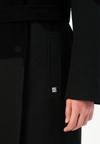 Primo piano di una mano accanto a un cappotto nero con un passante per cintura in tessuto che mostra un piccolo logo bianco "HUGO".