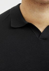 Juodas polo marškinėliai, pagaminti iš tekstūruoto mezginio audinio, su klasikinio tipo apykakle ir V formos iškirpte. Matomi subtilūs siuvimo akcentai.