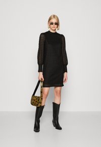 Vestido preto com tecido texturizado, mangas longas transparentes e decote redondo. Combinado com botas pretas até o joelho e uma mala de ombro estampada.