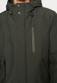 Veste isolante vert foncé avec une poche zippée, des boutons-pression et des cordons de capuche réglables. Tissu à texture lisse et design minimaliste.
