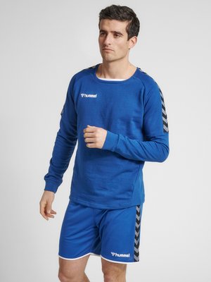 Blå langærmet skjorte med rund halsudskæring og sorte stribede detaljer på skuldrene, parret med matchende blå shorts med hvide kanter.