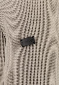 Texturiertes beiges Gewebe mit einem Rautenmuster, das ein kleines schwarzes Etikett mit der Aufschrift "KEY LARGO" zeigt, befestigt mit silberfarbenem Zubehör.