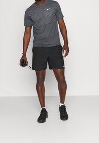 Szara, krótka koszulka sportowa Nike z teksturowanym wzorem, czarne spodenki i czarne trampki. Trzyma przezroczystą butelkę wody.