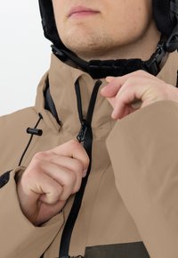Giacca beige con zip frontale e dettagli neri. Il colletto presenta un rivestimento morbido e sono visibili dei cordini nel cappuccio.