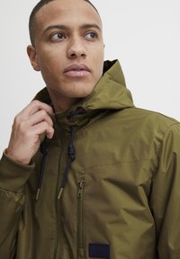 Olive green hooded jacket with zipper and drawstring details, featuring a chest pocket and navy accents for a contrast. Smooth fabric texture.