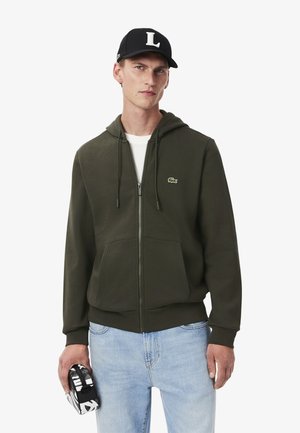 Sudadera con cremallera color verde oliva, con capucha ajustable, bolsillos frontales y un pequeño logo. Combinada con jeans de color azul claro y una gorra negra.