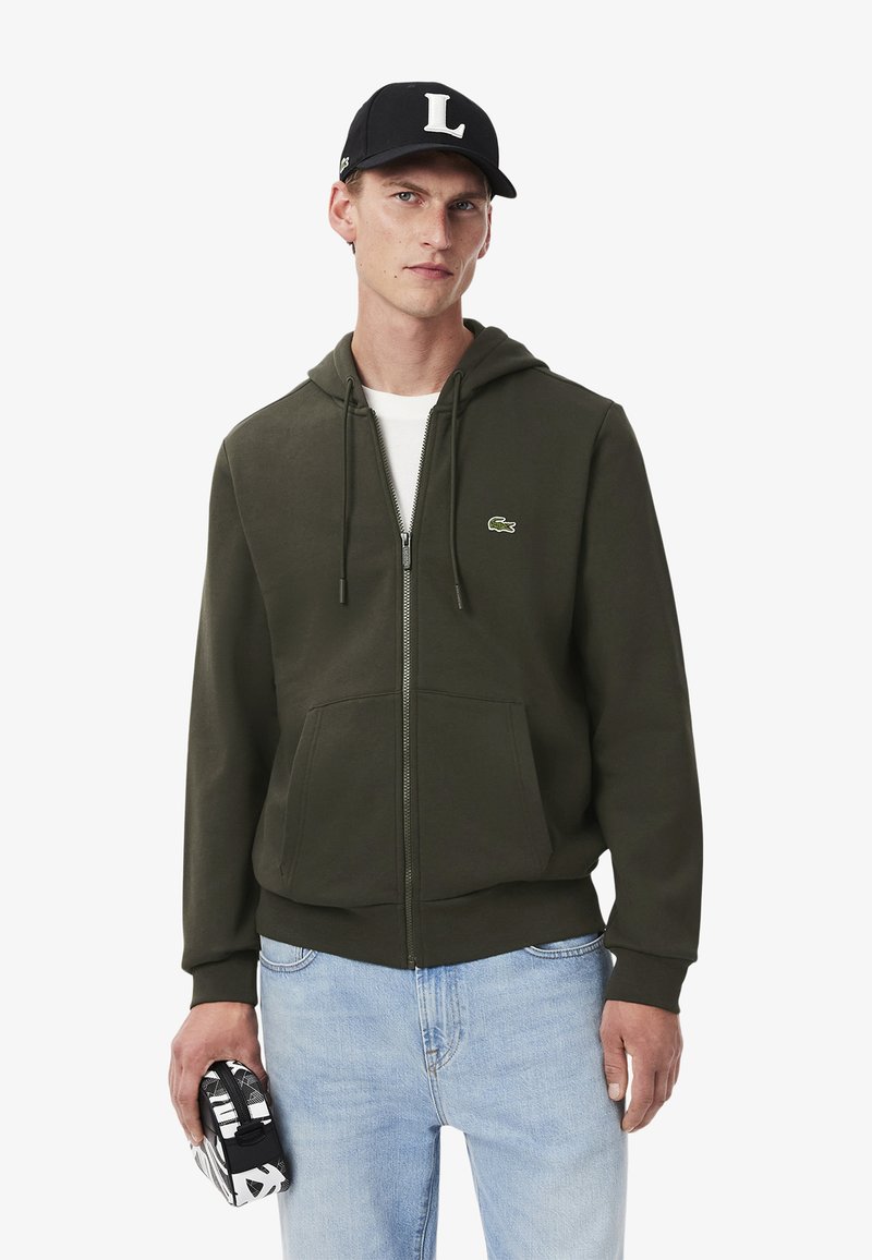 Veste à capuche zippée olive avec un cordon de serrage, poches avant et un petit logo. Portée avec un jean bleu clair et une casquette noire.
