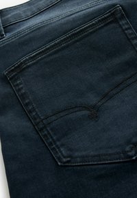 Gros plan de la poche arrière d’un jean en denim bleu foncé avec des coutures visibles et un détail subtil de surpiqûre courbée sur la poche.