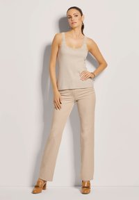 Beige Top mit Spitzenbesatz, kombiniert mit hellbeigen Schlaghosen. Das Model trägt braune High Heels. Vor einem neutralen Hintergrund.