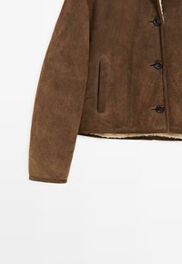 Bruine suèdeジャケット met een knoopsluiting aan de voorkant, zijzakken en een textuurafwerking. De binnenkant heeft een zachte, lichtgekleurde voering.