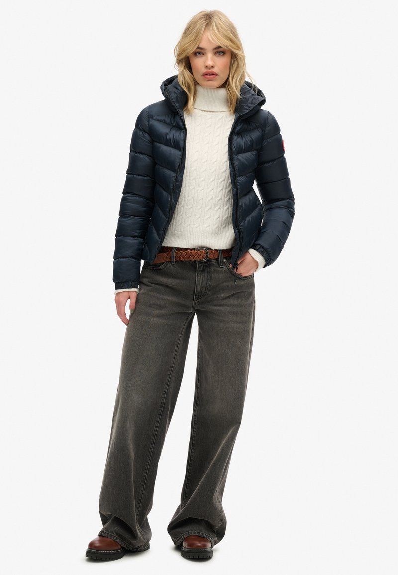 Marine pufferjack met capuchon, met een gequiltde textuur, gedragen over een crèmekleurige kabelgebreide trui; gecombineerd met brede grijze jeans en bruine laarzen.