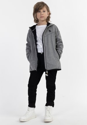 Giacca grigia con zip, interno nero, tasche frontali e dettaglio del logo, abbinata a pantaloni neri e sneaker alte bianche.