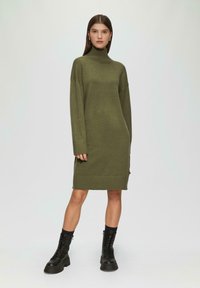 Olivegrünes Strickkleid mit hohem Kragen, oversized Passform, gerippten Bündchen und Saum. Kombiniert mit schwarzen Schnürkampfschuhen.