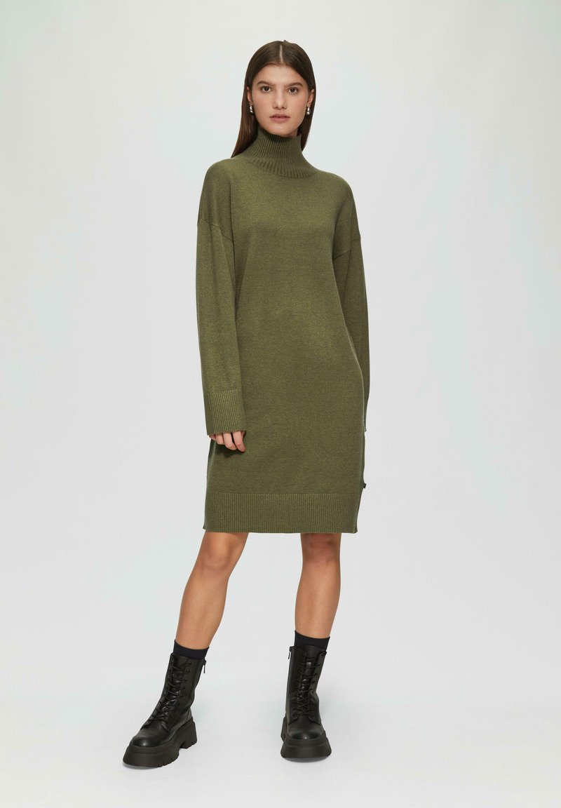 Olivegrünes Strickkleid mit hohem Kragen, oversized Passform, gerippten Bündchen und Saum. Kombiniert mit schwarzen Schnürkampfschuhen.