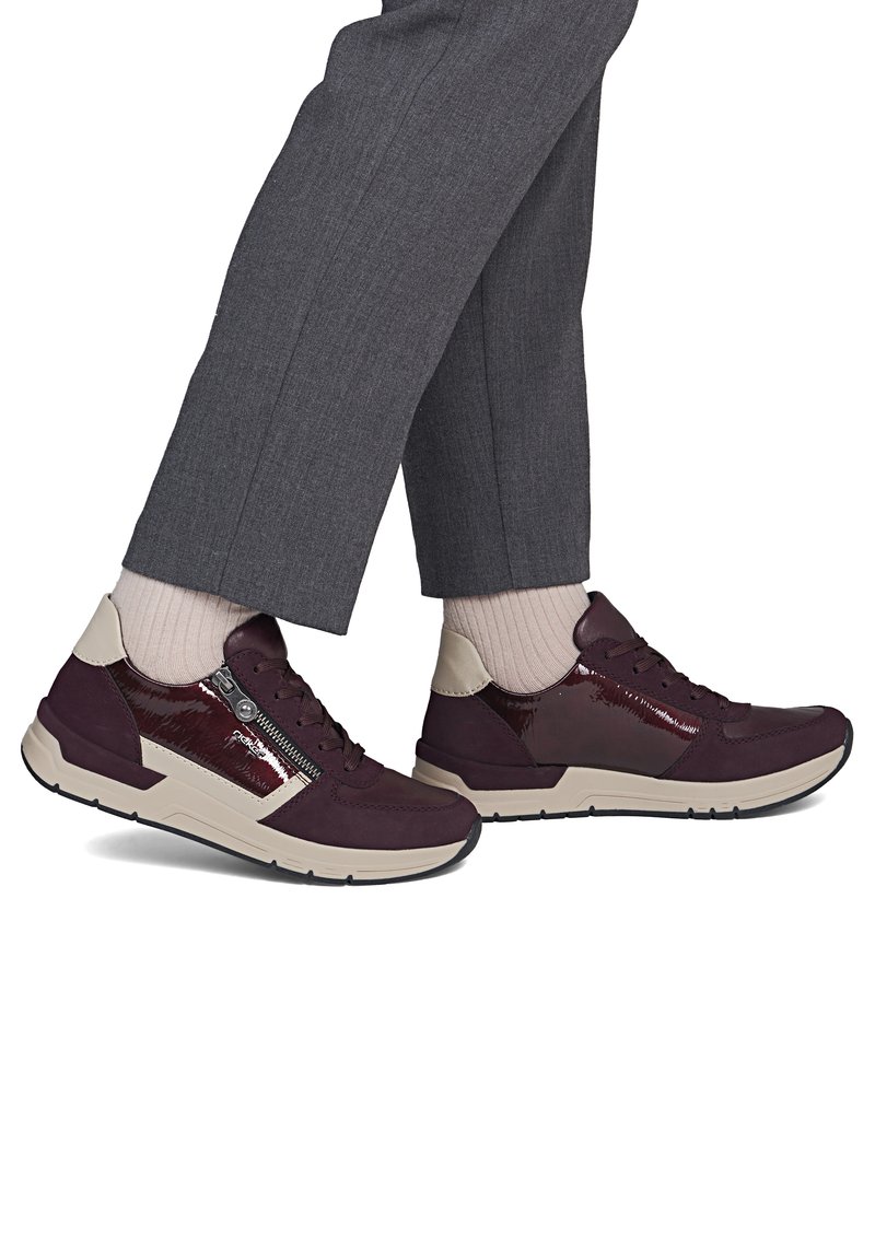 Personne portant un pantalon gris foncé, des chaussettes beiges et des baskets violet foncé avec semelles beiges et détail de fermeture éclair sur le côté, debout, les pieds croisés.