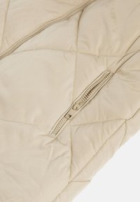 Chaqueta acolchada beige con un patrón de acolchado diagonal, un bolsillo con cremallera y un acabado suave y brillante.