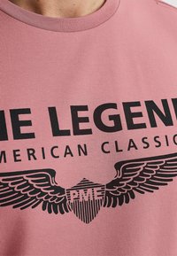 Camiseta de algodón rosa claro con texto negro en negrita "LA LEYENDA" y "CLÁSICO AMERICANO", con gráficos de alas y escudo.