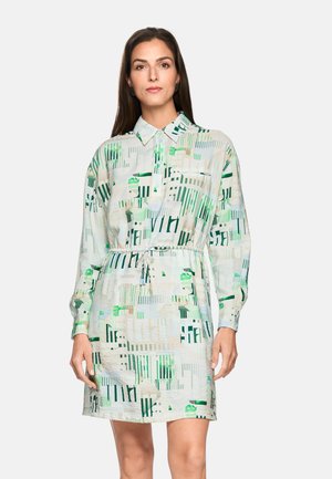 Woman wearing a long-sleeve, collared shirt dress with abstract green and beige geometric patterns and a drawstring waist, standing against a white background.