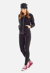Black athletic jacket with zipper and side pockets, paired with black leggings. Accented with a black beanie and red sneakers.