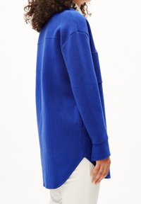 Blå fleece-tunika med rundad nederkant, långa ärmar och sidofickor. Har en mjuk textur och en avslappnad passform, visad i profil.