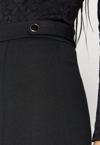 High-waisted black pants made of stretch fabric with a decorative button and flat front, featuring smooth texture and subtle stitching details.
