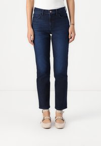 Jeans de ganga azul escura com perna reta, cintura alta e desbotamento sutil. Combinados com sapatos de tira no tornozelo de cor clara.