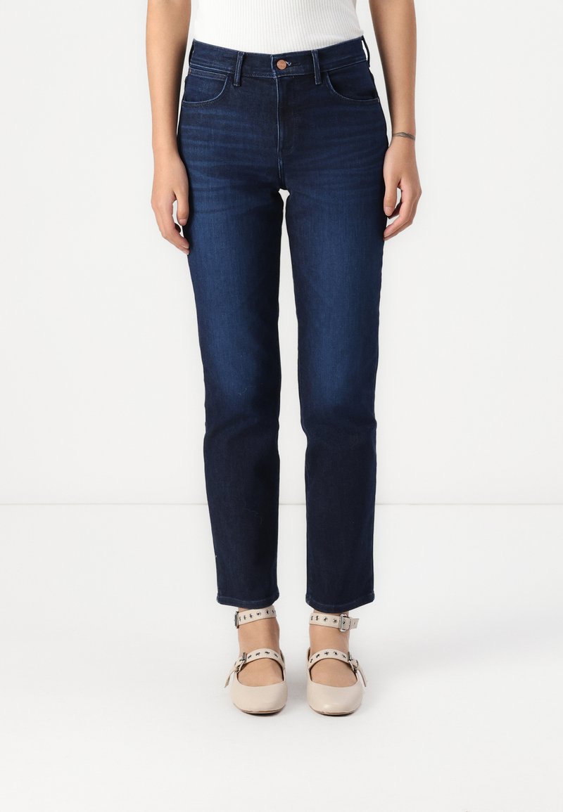 Jeans de ganga azul escura com perna reta, cintura alta e desbotamento sutil. Combinados com sapatos de tira no tornozelo de cor clara.