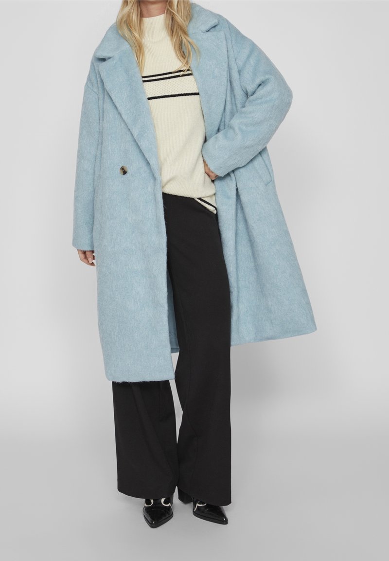Femme portant un manteau oversize bleu clair sur un pull crème à rayures noires et un pantalon noir à jambes larges, debout avec une main dans la poche.