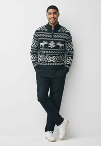 Schwarzer und weiß gemusterter Pullover mit Elch- und Baumdesigns, kombiniert mit dunklen Hosen und weißen Sneakers. Das Modell posiert vor einem schlichten Hintergrund.