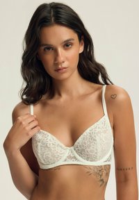 Biała koronkowa biustonosz o strukturalnym wzorze, z cienkimi ramiączkami, kwiatowymi wzorami i gładką taśmą pod biustem. Dodatki to kokarda na środku.