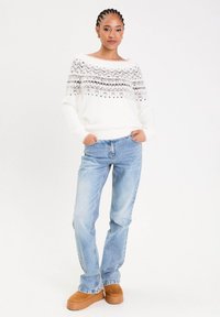 Femme à cheveux tressés portant un pull blanc à motifs, un jean bleu clair et des chaussures à plateforme marron, se tenant les mains dans les poches.