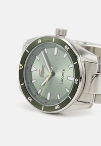 Ceas din oțel inoxidabil cu bezel verde și cadran verde deschis. Dispune de markeri albi pentru ore, un ac de secundă galben și logo-ul Lacoste.